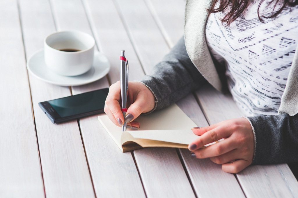 一位女子拿著筆在筆記本紀錄，桌子旁邊放咖啡與手機